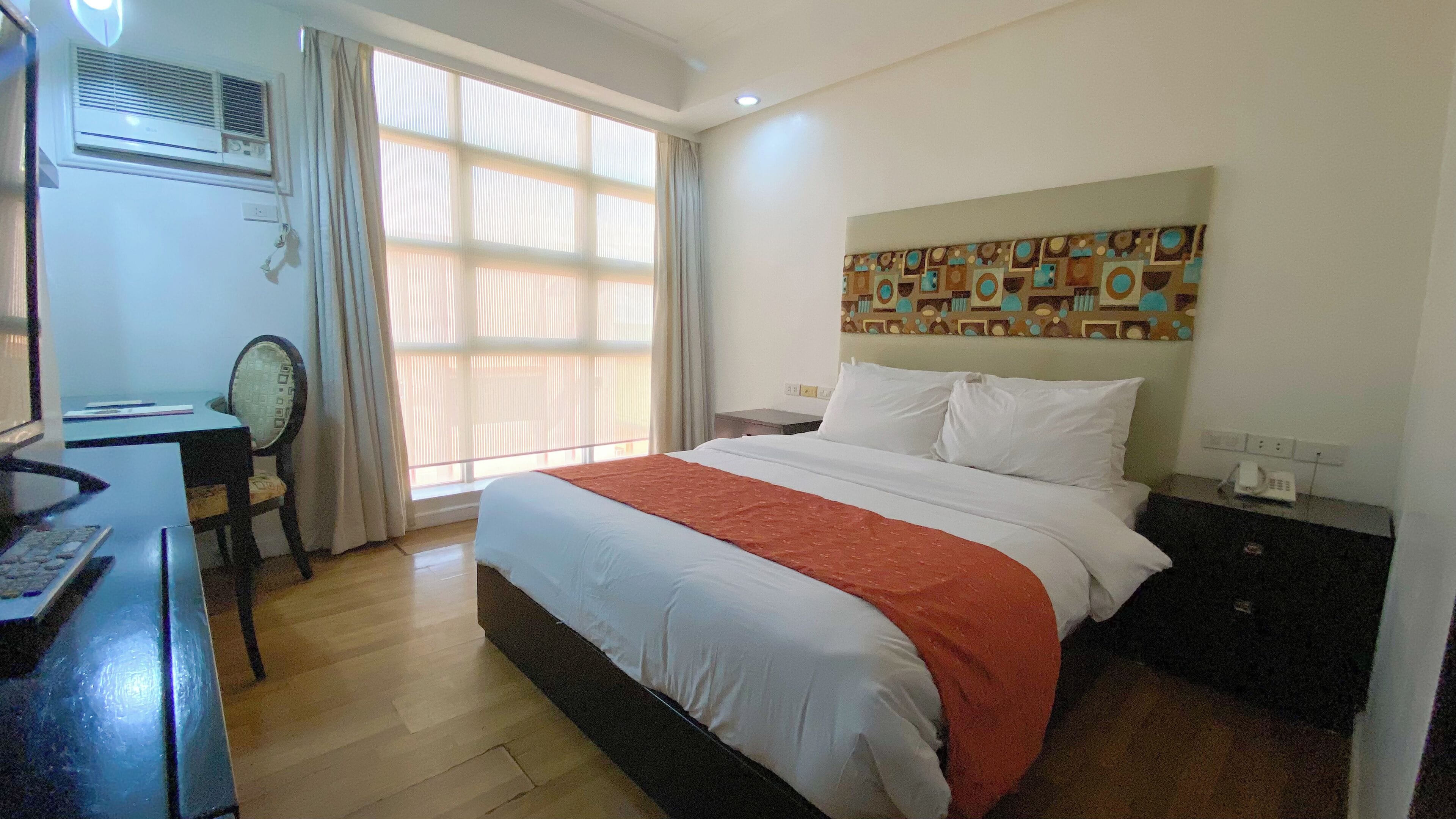 Ein Hotelzimmer mit Bett, Schreibtisch mit Stuhl, Fenster mit Jalousien, Wand-Klimaanlage und einer Wandverkleidung.