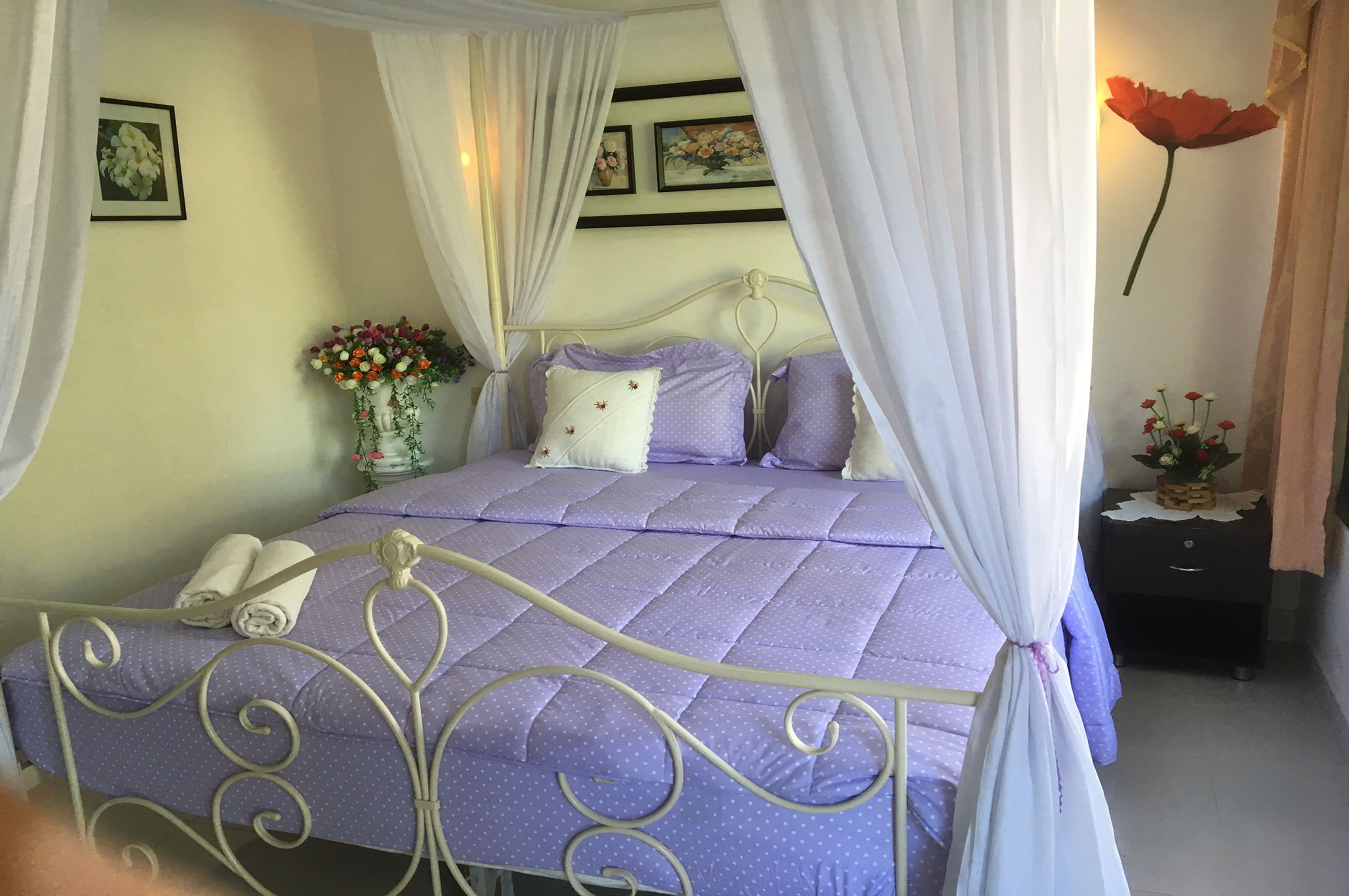 Una cama bien hecha con una colcha morada y cortinas blancas, una mesita de noche con un florero y un adorno floral en la pared.