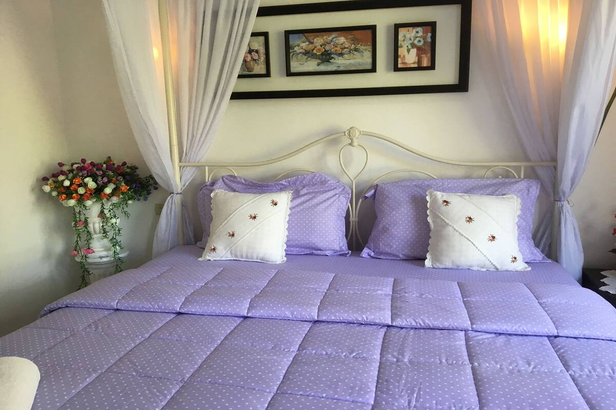 A neatly made bed with a purple quilt and white pillows, a white metal headboard, and a wall with framed artwork.