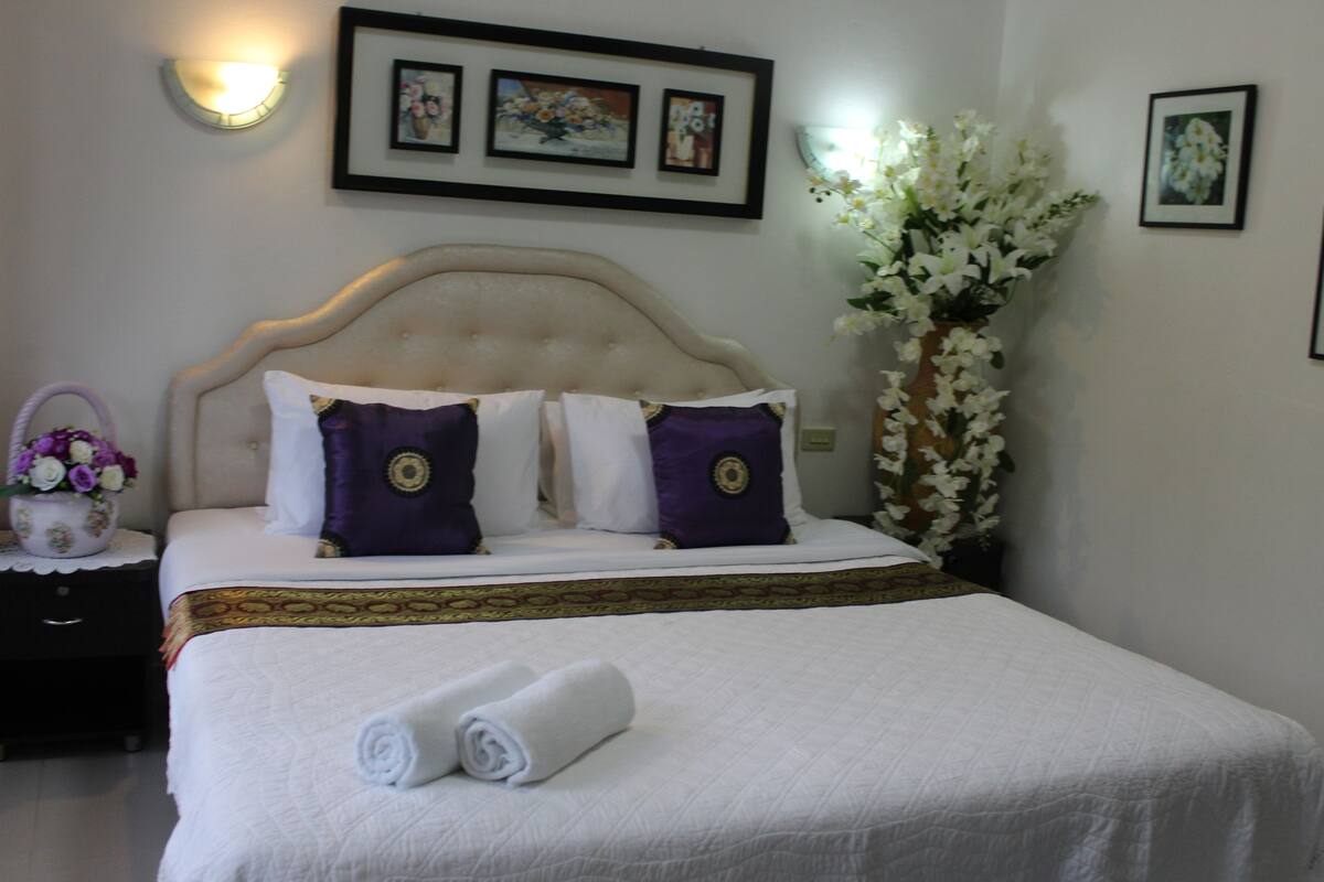 A neatly made bed with a white duvet, two purple pillows, and a rolled white towel on the footboard.