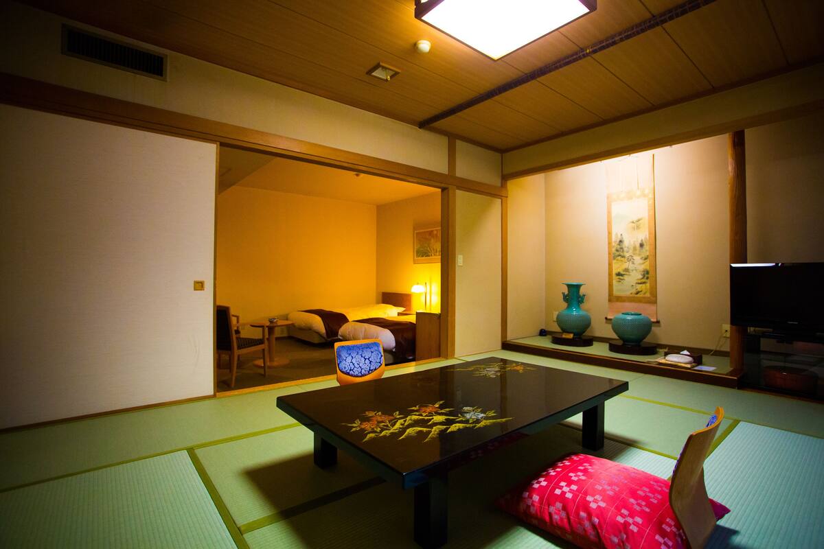 A traditional Japanese room with tatami flooring, a low table, a red cushion, and a television.