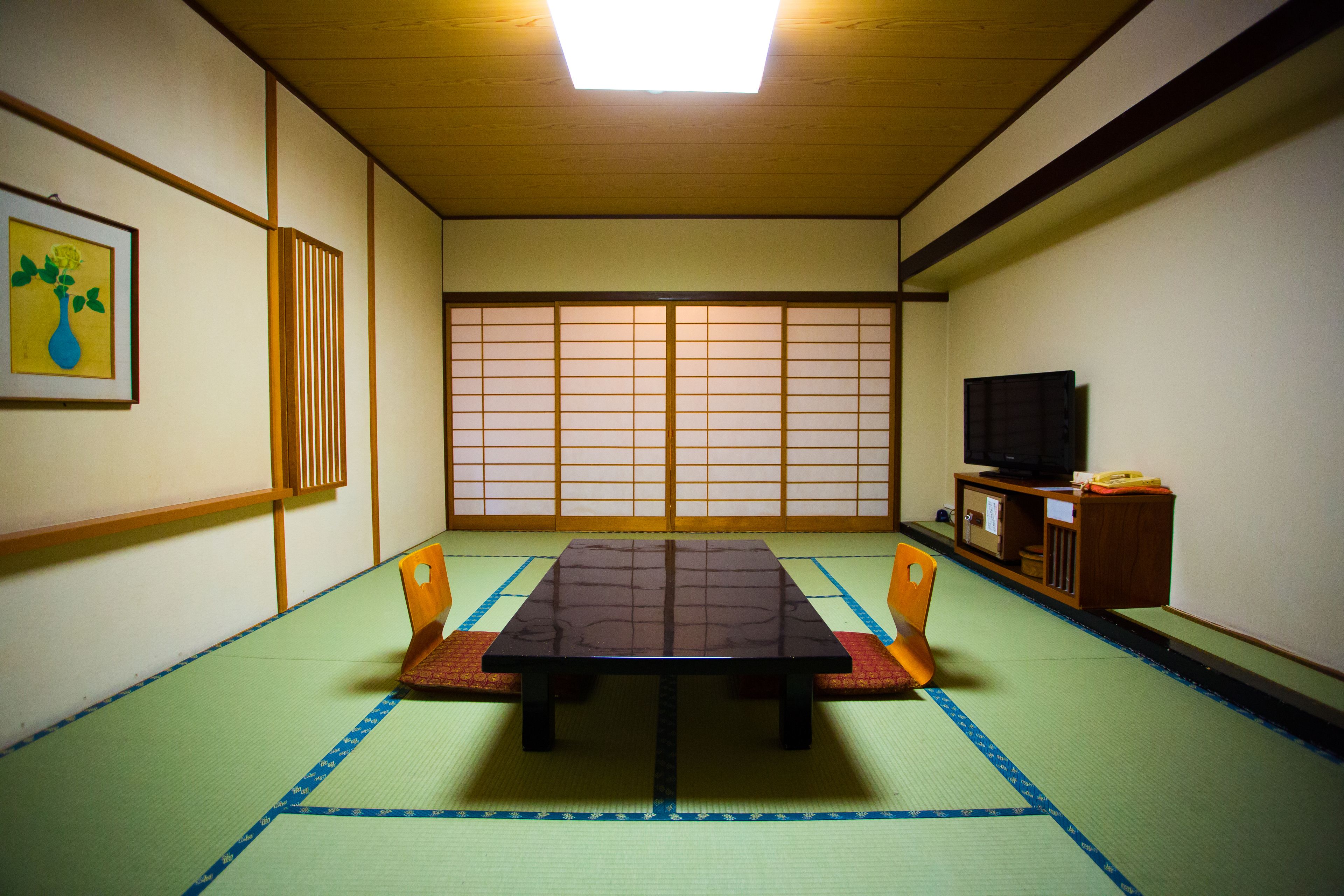 Ein Raum mit Tatami-Matten, einem niedrigen Tisch, Holzstühlen, einem Fernseher und einem Shoji-Bildschirm.