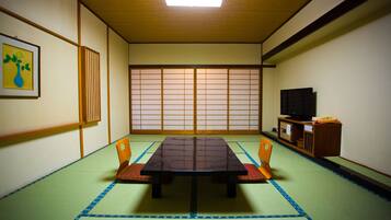 Ein Raum mit Tatami-Matten, einem niedrigen Tisch, Holzstühlen, einem Fernseher und einem Shoji-Bildschirm.