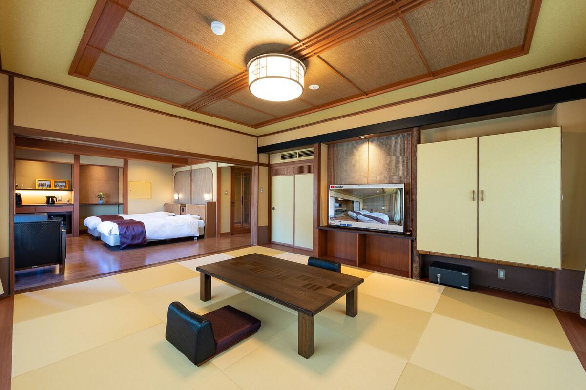 A hotel room with a large bed, a dining table, a small kitchen area, and a wardrobe.