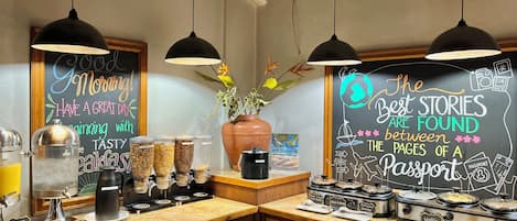 A breakfast bar with coffee machines, a fruit juice dispenser, and a chalkboard with motivational messages.