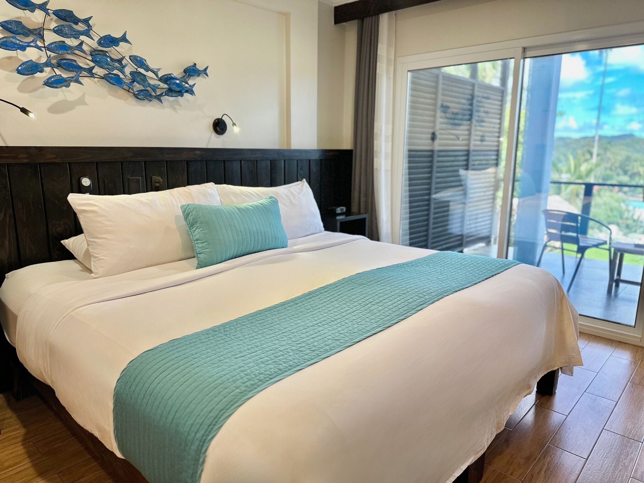 Habitación de hotel con cama, decoración de peces en la pared, vistas al exterior y balcón con sillas.