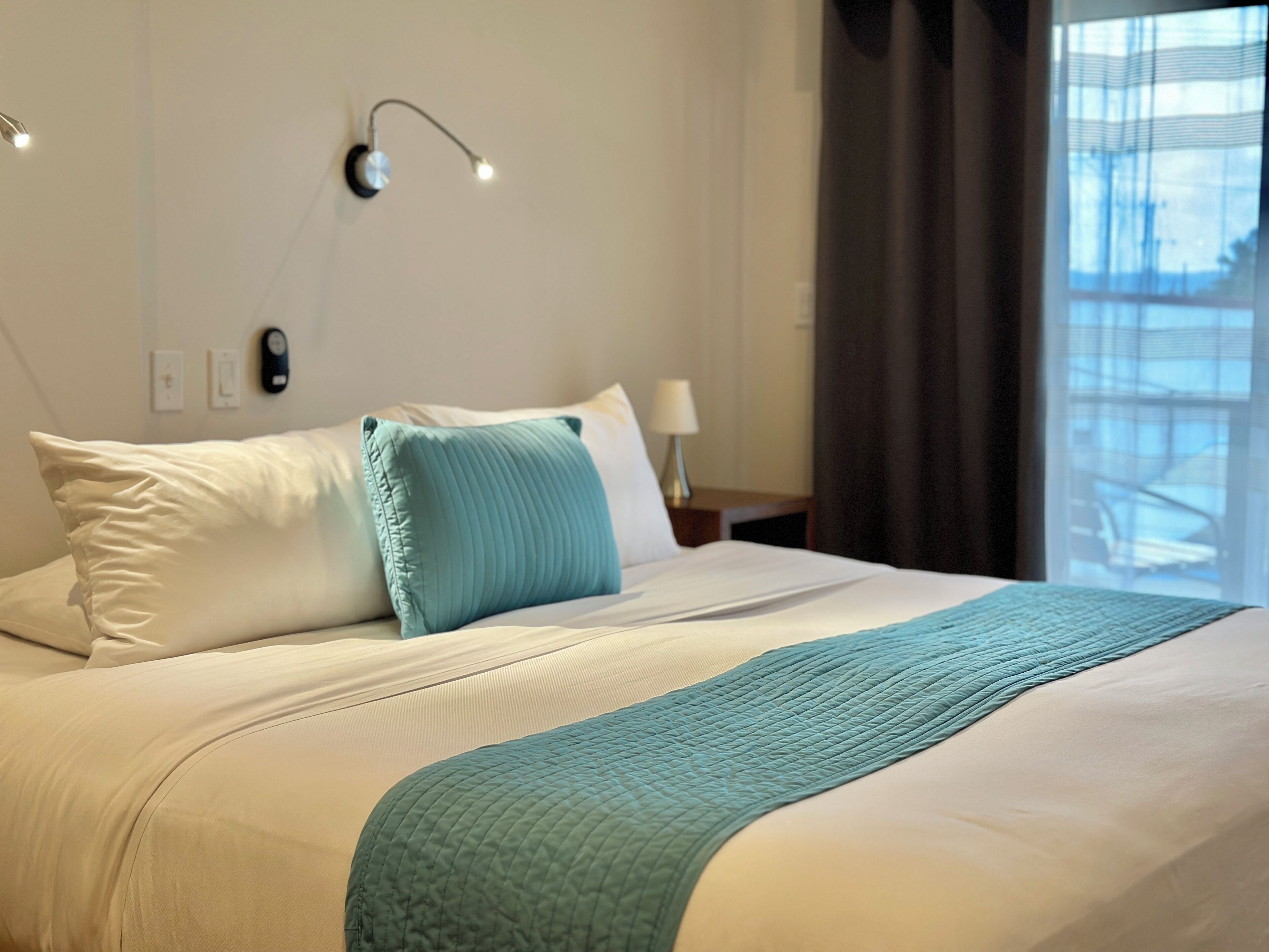 Ein ordentlich bezogenes Bett mit einem türkisfarbenen Kopfkissen und einer gesteppten Decke. Ein Nachttisch mit Lampe und einer Wandleuchte sind vorhanden.