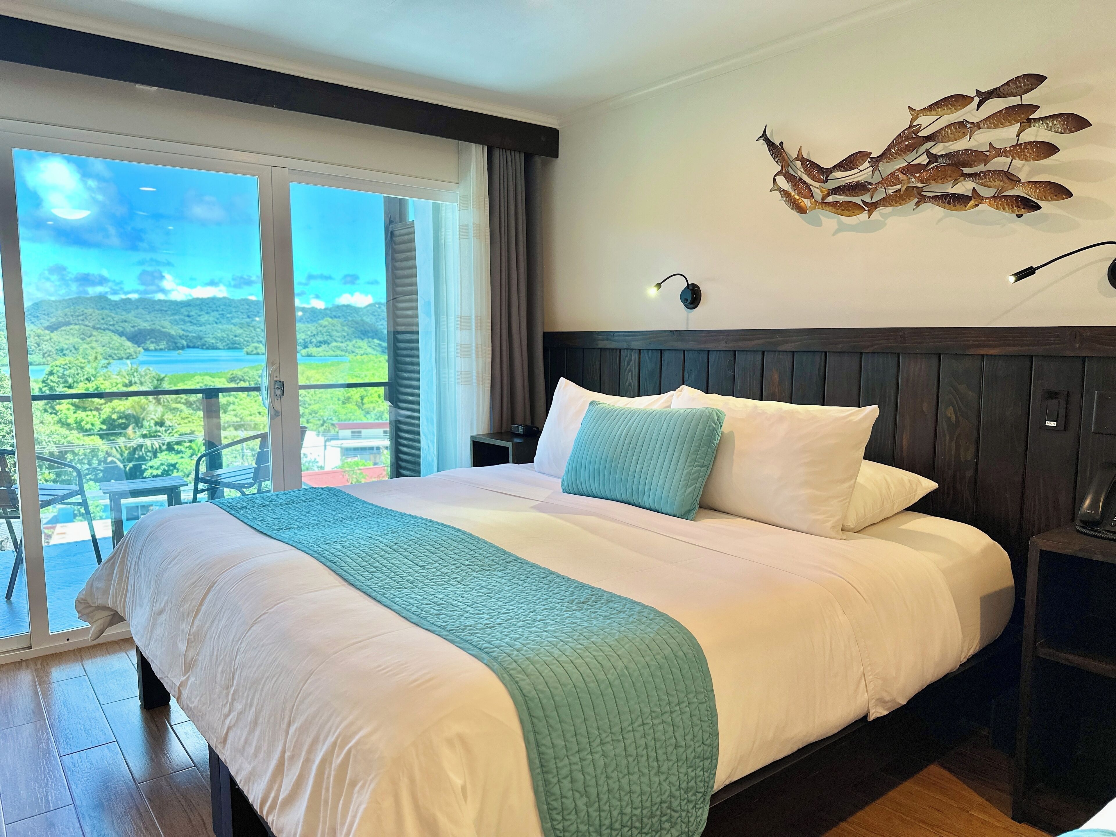A hotel room with a large bed, a view of a lush landscape through a balcony, and a decorative wall piece.