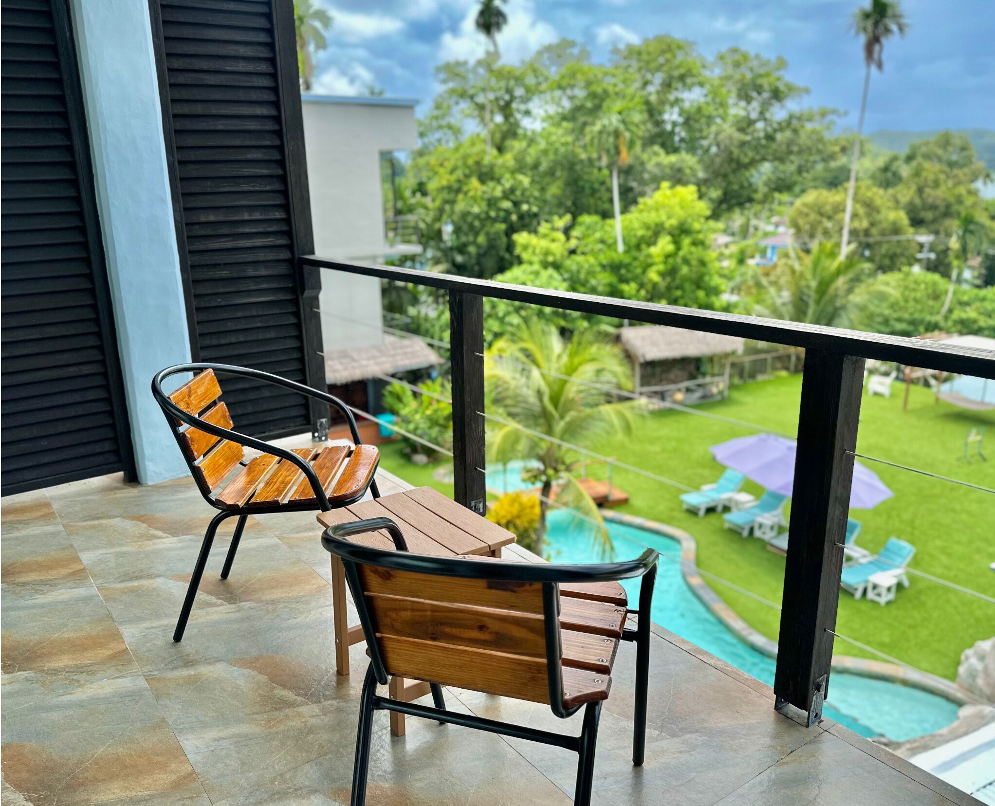 Un balcón con dos sillas y una mesa de madera, con vista a una piscina y abundante vegetación.