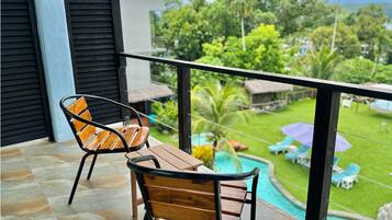 A balcony with two wooden chairs and a table, overlooking a swimming pool and lush greenery.