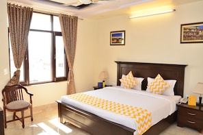 A hotel room with a large bed, a wooden chair, a nightstand, and a window with curtains. - Raj Regency (Jaipur)