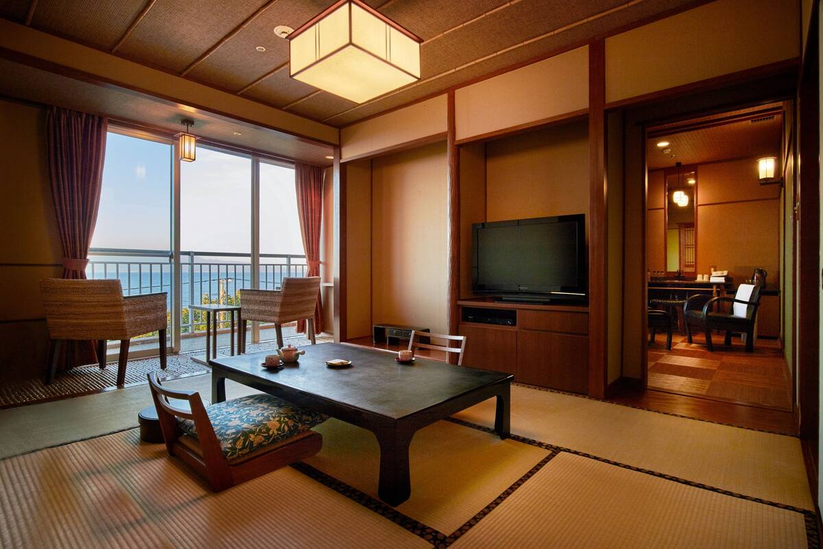 Een traditionele Japanse kamer met tatami vloeren, een lage tafel, stoelen, een televisie en een balkon met uitzicht.