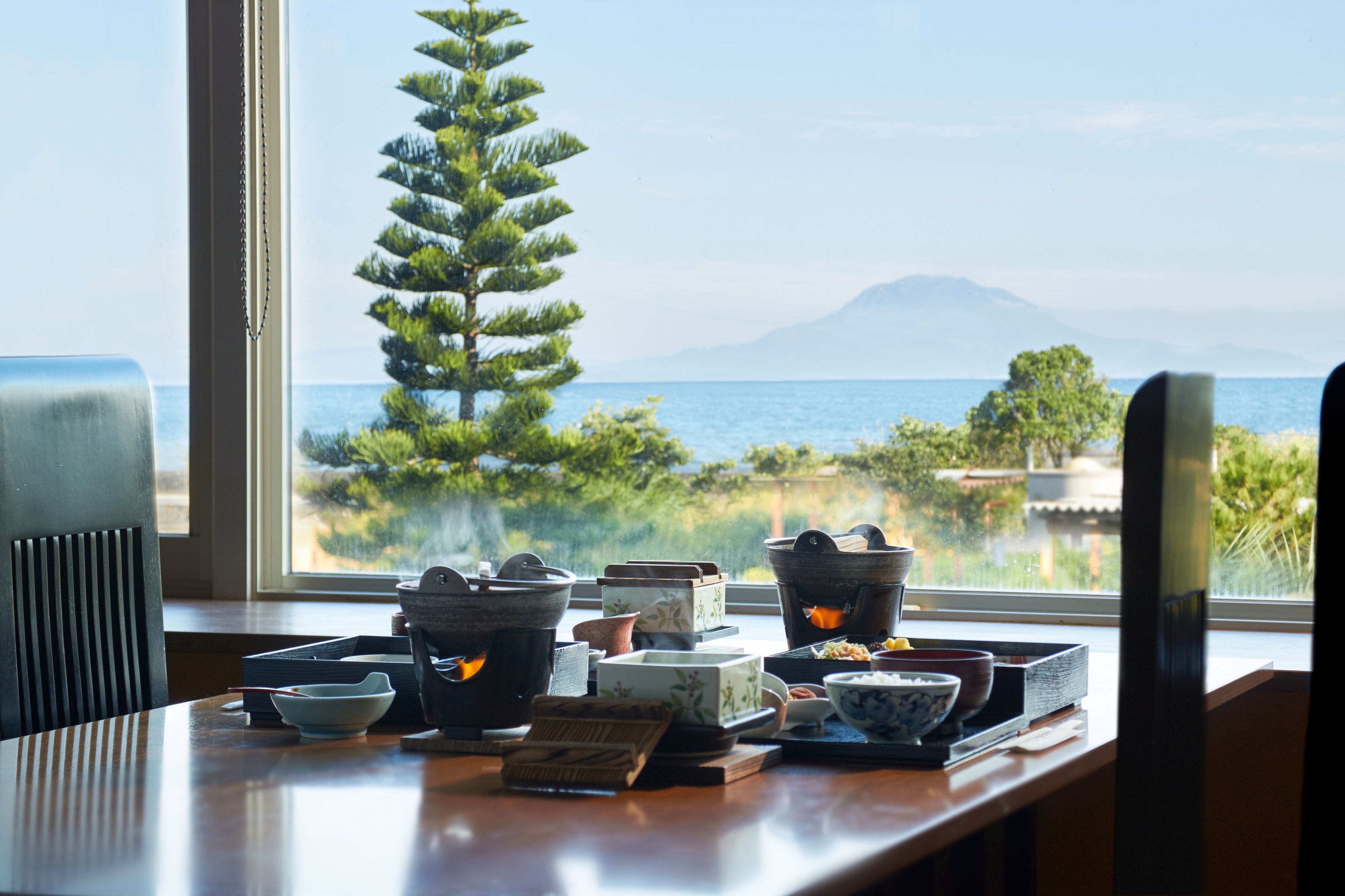 A table with various ceramic dishes and a pine tree by a window overlooking a body of water and a mountain.