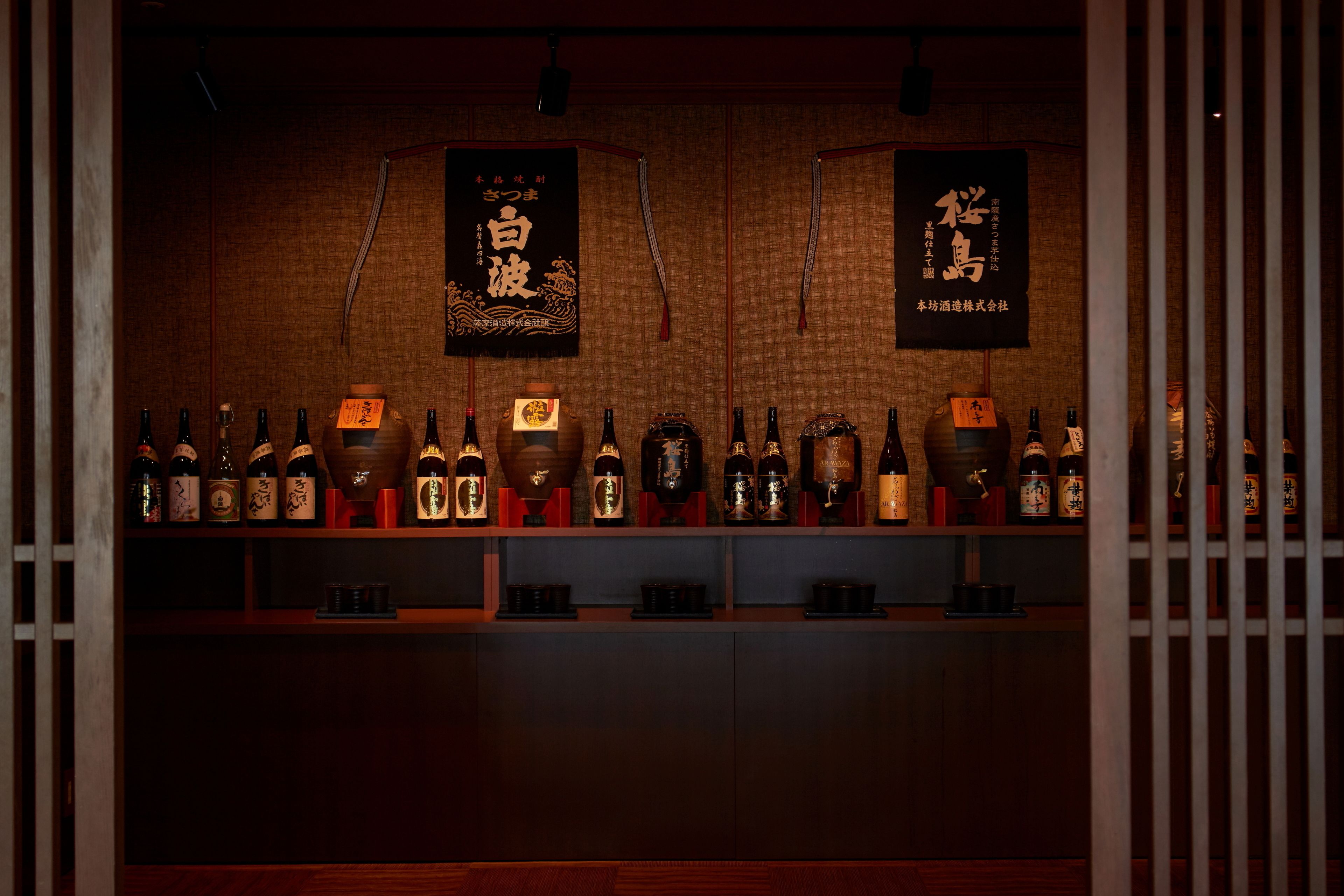 Zona de bar con estantes llenos de botellas y dos pancartas con caracteres japoneses.