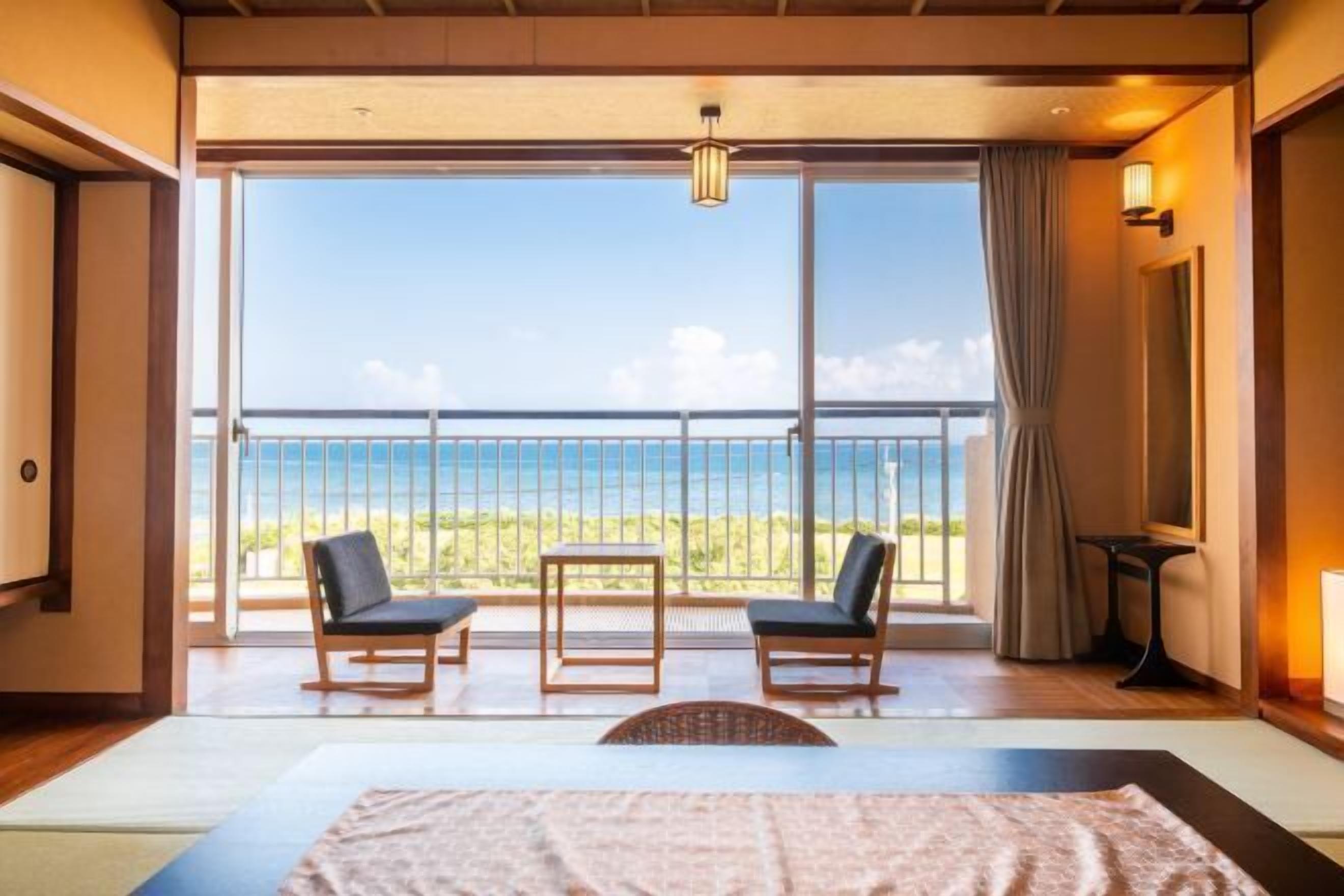 A hotel room with a large balcony, a bed, two chairs, a small table, and a view of the sea.
