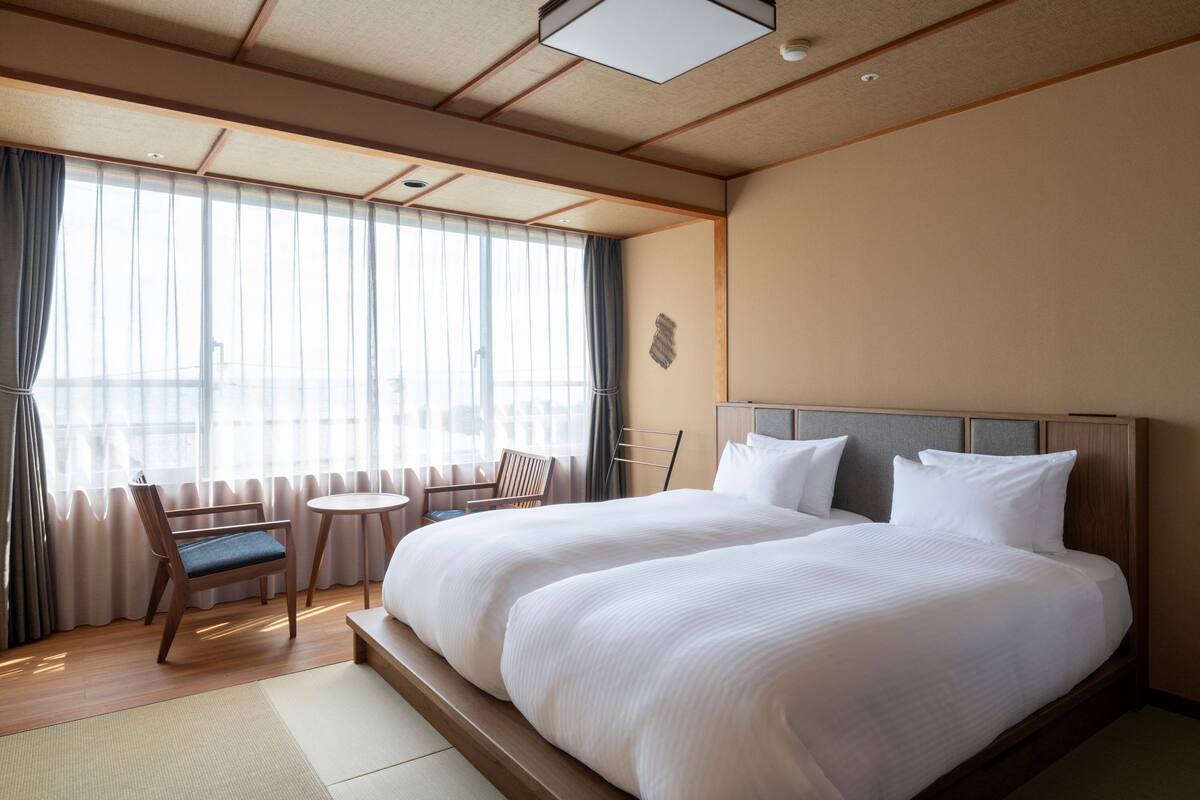 Een hotelkamer met een groot bed, een stoel, een klein tafeltje en uitzicht op buiten door grote ramen.