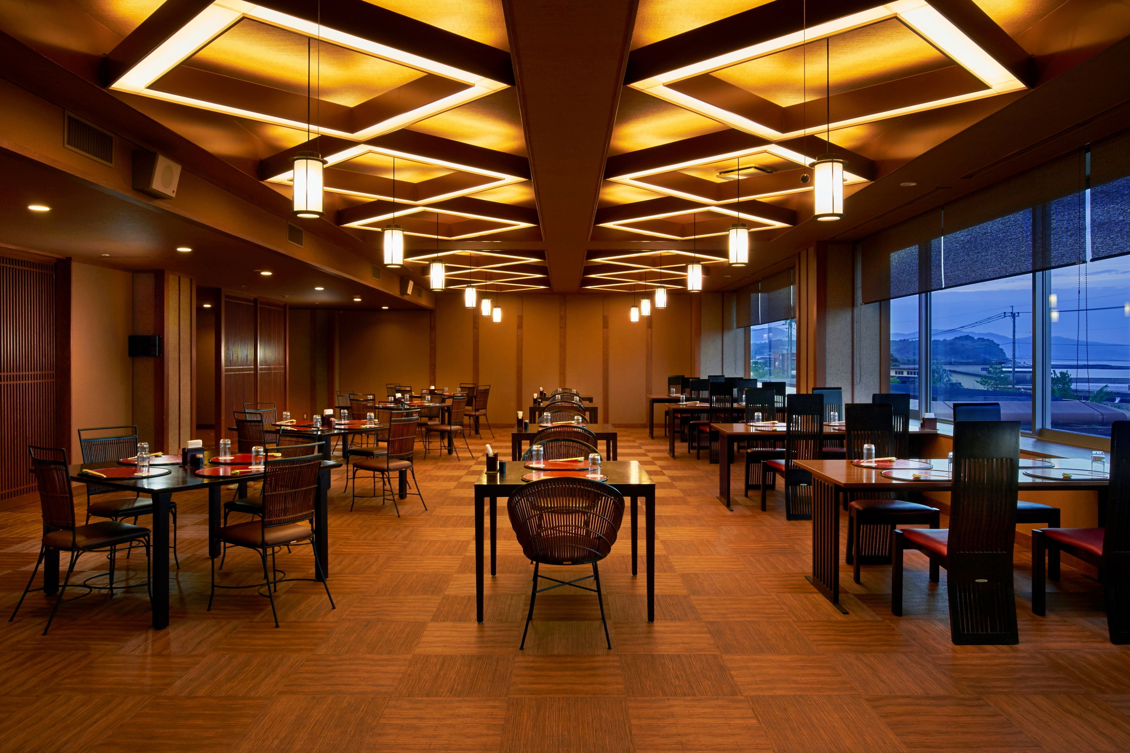 A spacious hotel dining area with multiple tables and chairs, large windows offering a view of the outside, and a modern ceiling design with recessed lighting.