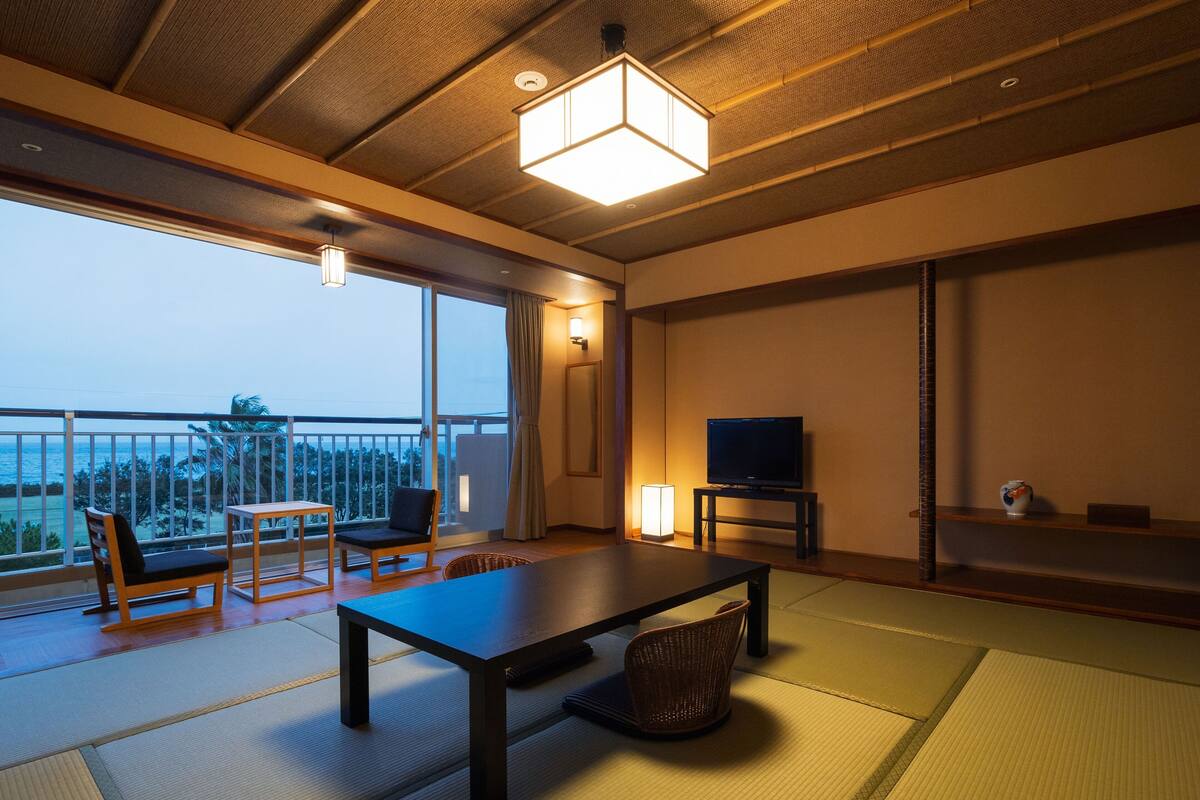 Een moderne woonkamer met tatami matten, een lage tafel, een bank, een televisie en een balkon met uitzicht op zee.