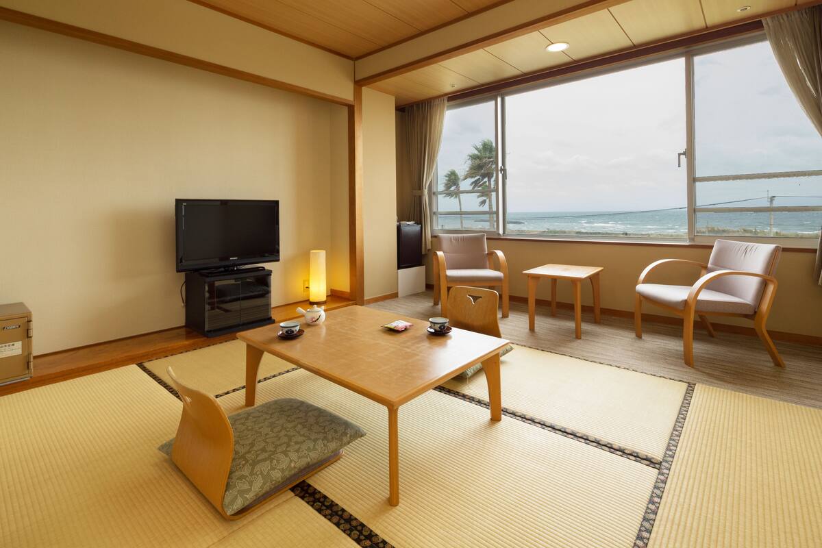 Een hotelkamer met tatami matten, een houten tafel, stoelen, een televisie en uitzicht op de oceaan.