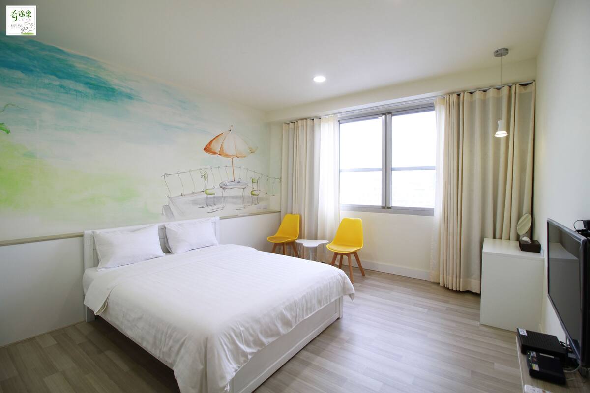 A hotel room with a large bed, two yellow chairs, a small table, a TV, and a window with curtains.