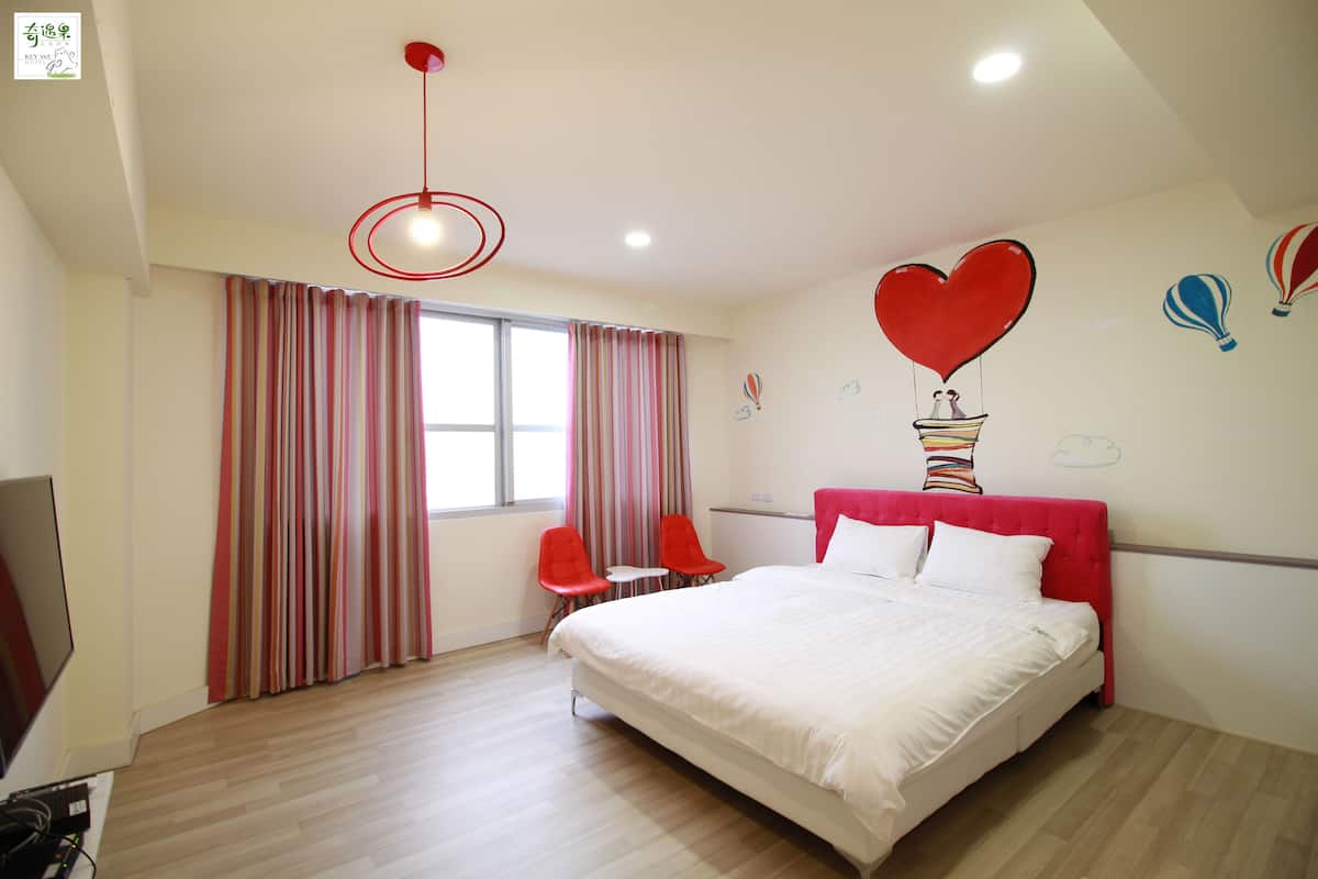 A hotel room with a large bed, two red chairs, a small table, a TV, and a window with curtains.