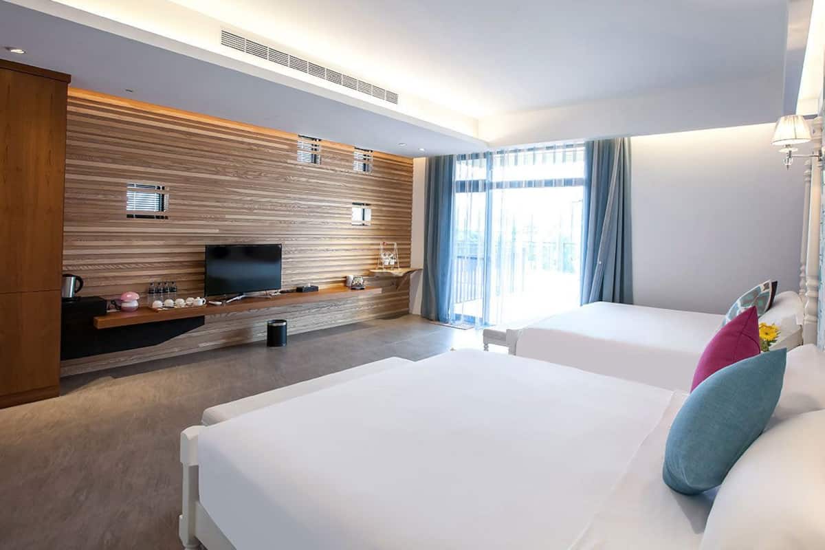 A modern hotel room with a wooden accent wall, a flat-screen TV, a bedside table with a lamp, and a large window with curtains.