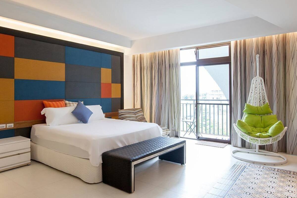 A modern hotel room with a large bed, a bedside table, a bench, a hanging chair, and a view of the outdoors through a large window.