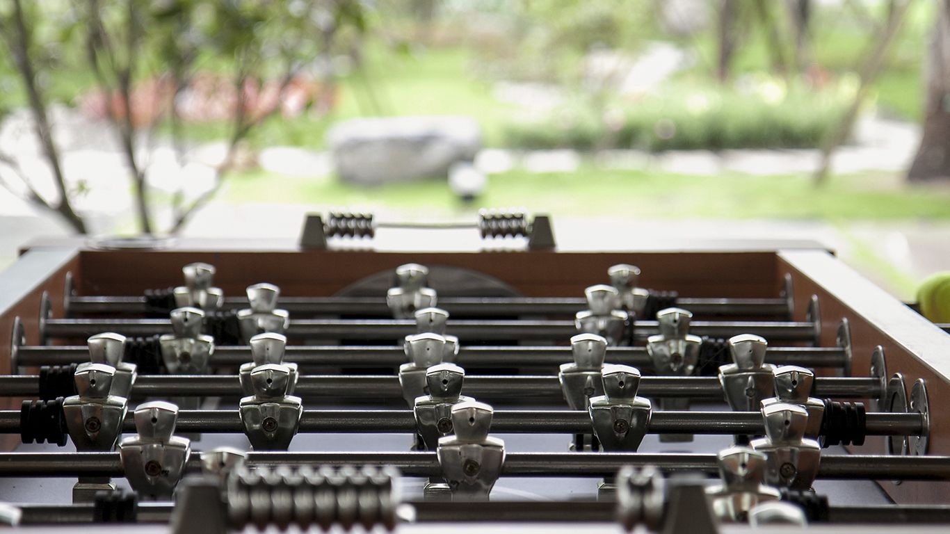 A close-up of a foosball table with metal rods and figures, set against a blurred outdoor background.