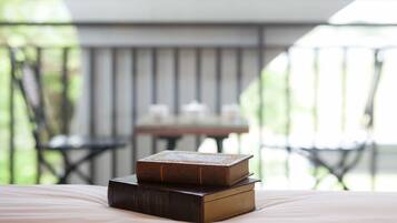 A stack of books on a bed in a room with a view of a balcony and outdoor furniture.