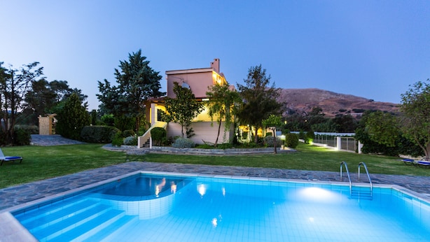 A swimming pool with steps leading into it, surrounded by a well-maintained lawn and a house with a lighted exterior.