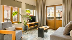Een moderne woonkamer met een bank, een salontafel en een televisie op een houten plank. Er is een groot raam met gordijnen, een balkon en een plant.