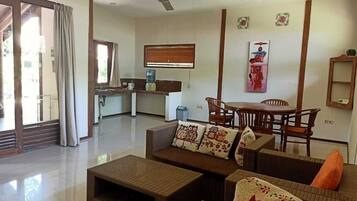 A living room with a sofa set, a dining table, and chairs. There is a kitchen area in the background.