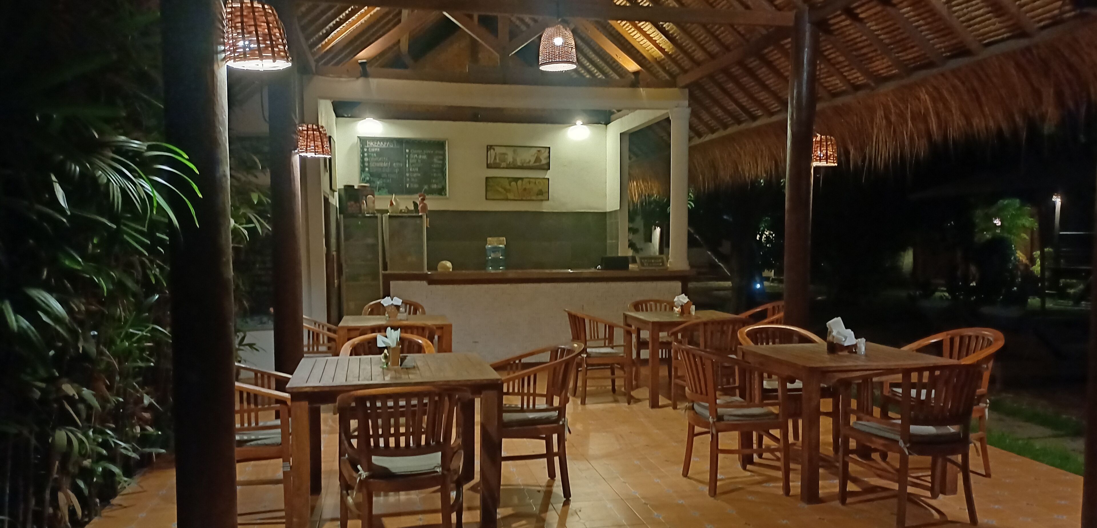 An outdoor dining area with wooden tables and chairs, a thatched roof, and a view of the surrounding greenery.
