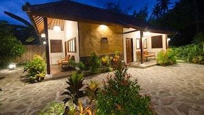 A two-story house with a tiled roof, a porch with wooden furniture, and a stone pathway leading to the entrance. - Si Pitung Village (Gili Air)
