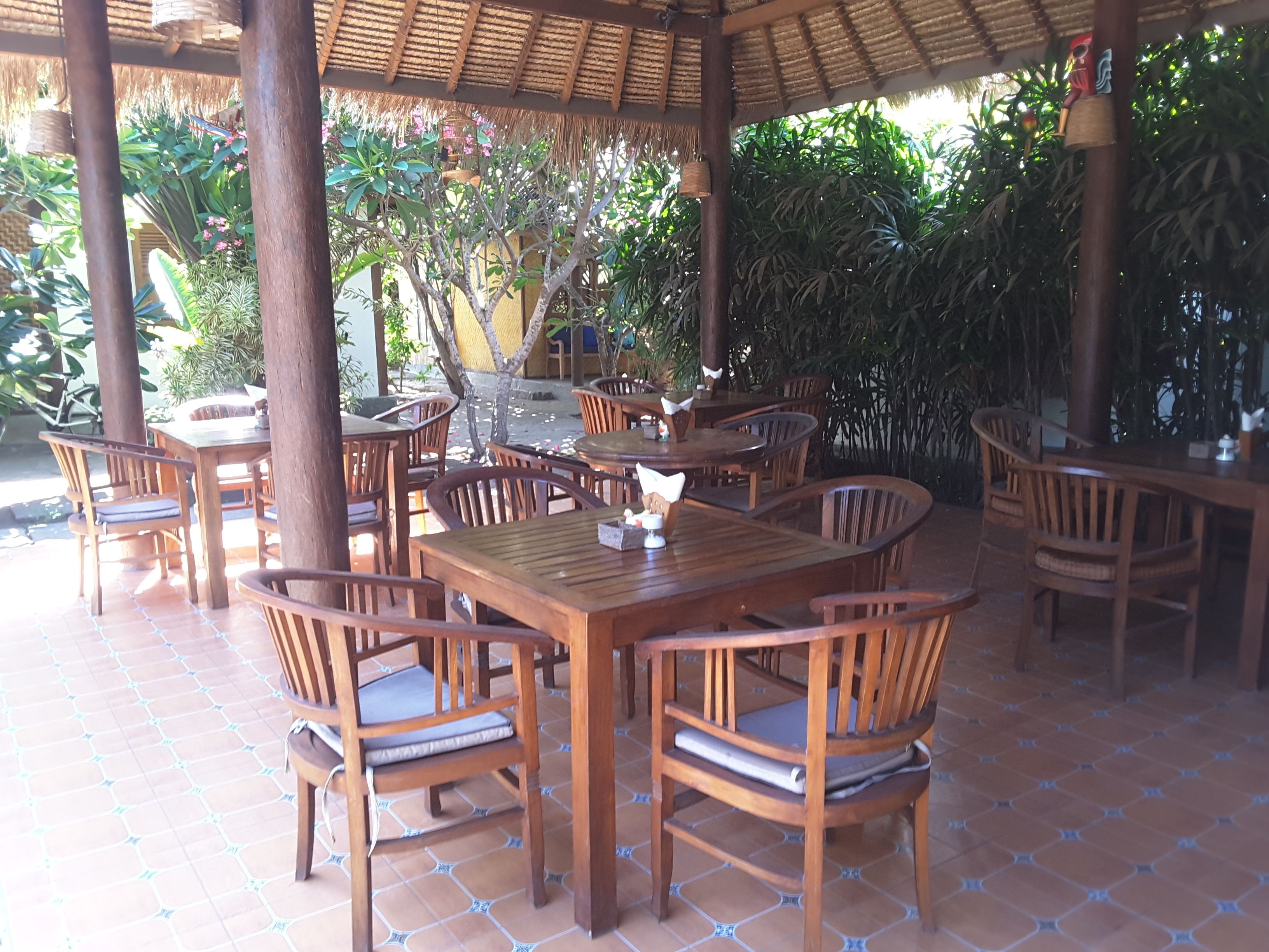 An outdoor dining area with wooden tables and chairs, surrounded by lush greenery and a thatched roof structure.