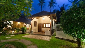 A cozy, well-lit cottage-style building with a tiled roof, surrounded by lush greenery and palm trees. - Si Pitung Village (Gili Air)