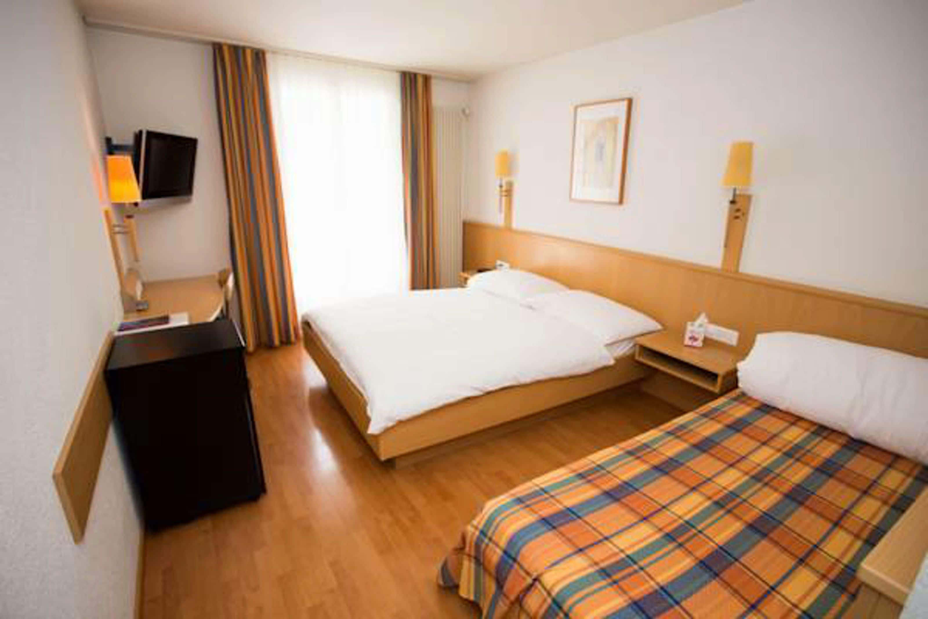 Una camera d'albergo con un letto, una scrivania con lampada, una TV e una finestra con tende.