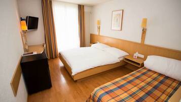 Una camera d'albergo con un letto, una scrivania con lampada, una TV e una finestra con tende.