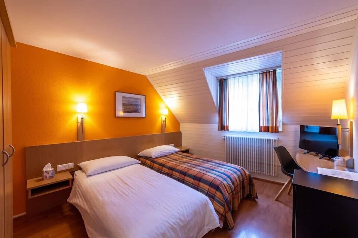 Ein Hotelzimmer mit zwei Betten, einem Schreibtisch mit Computer, einem Fenster mit Vorhängen und Wandlampen.