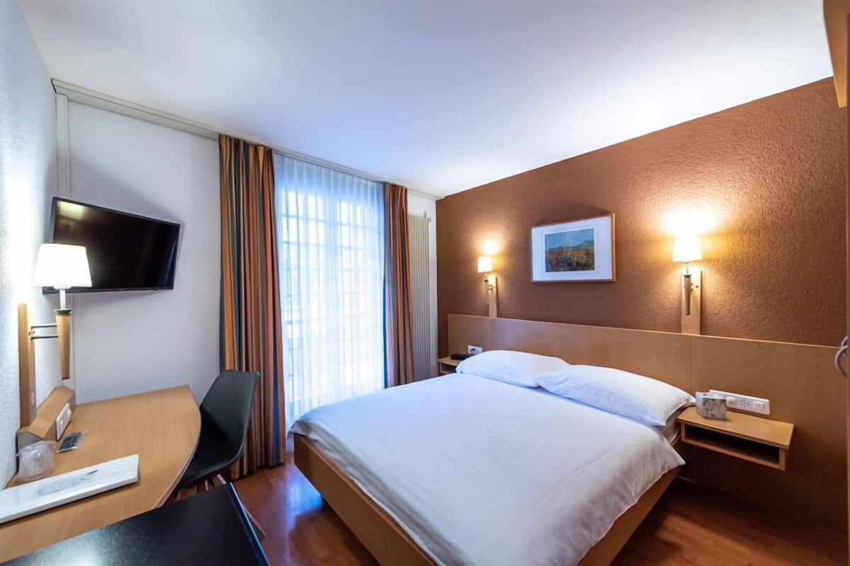 Ein Hotelzimmer mit Bett, Schreibtisch, Stuhl, Fernseher und einem Fenster mit Vorhängen.