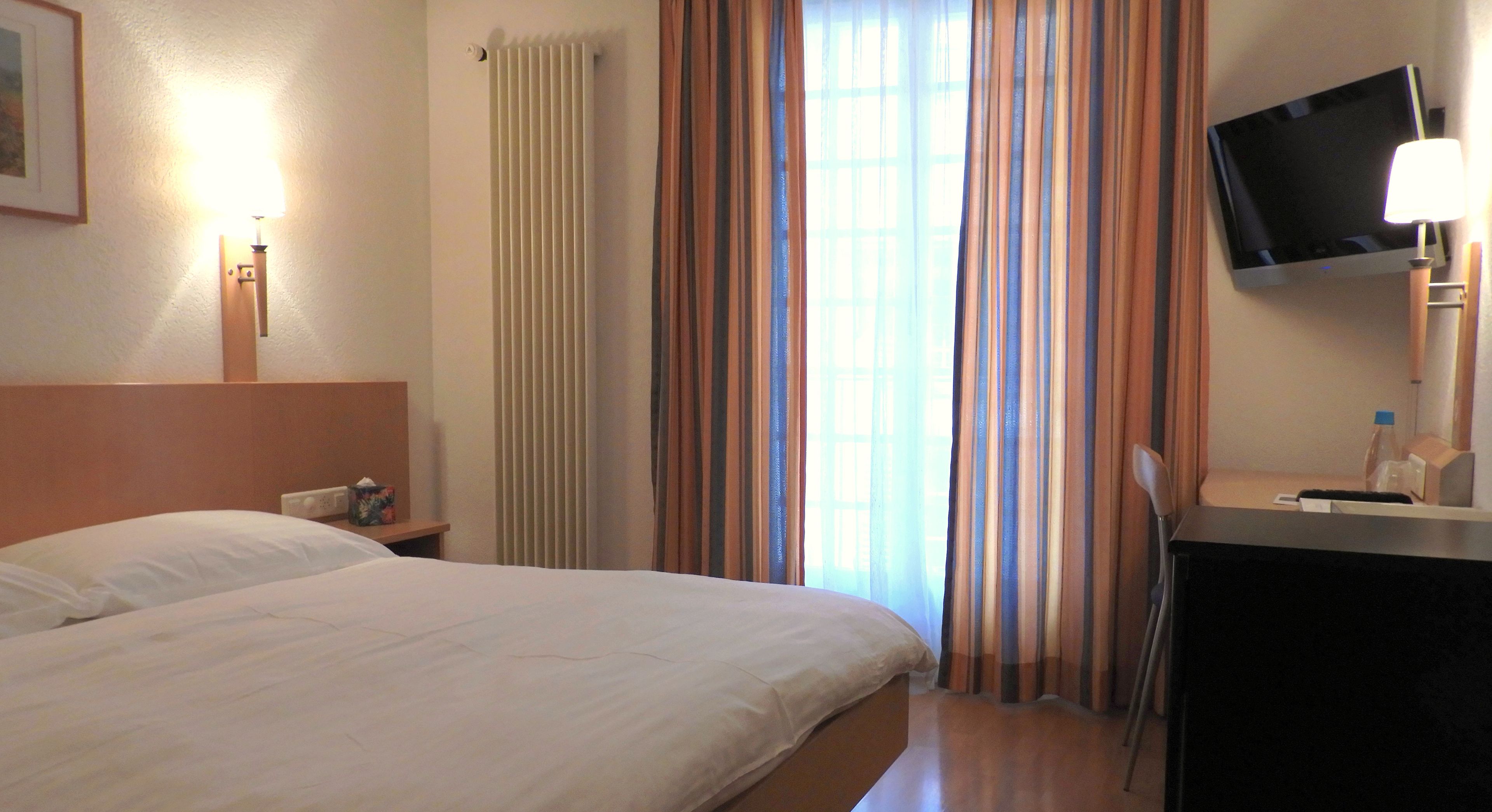 Habitación de hotel con cama, escritorio, silla, televisor y ventana con cortinas.