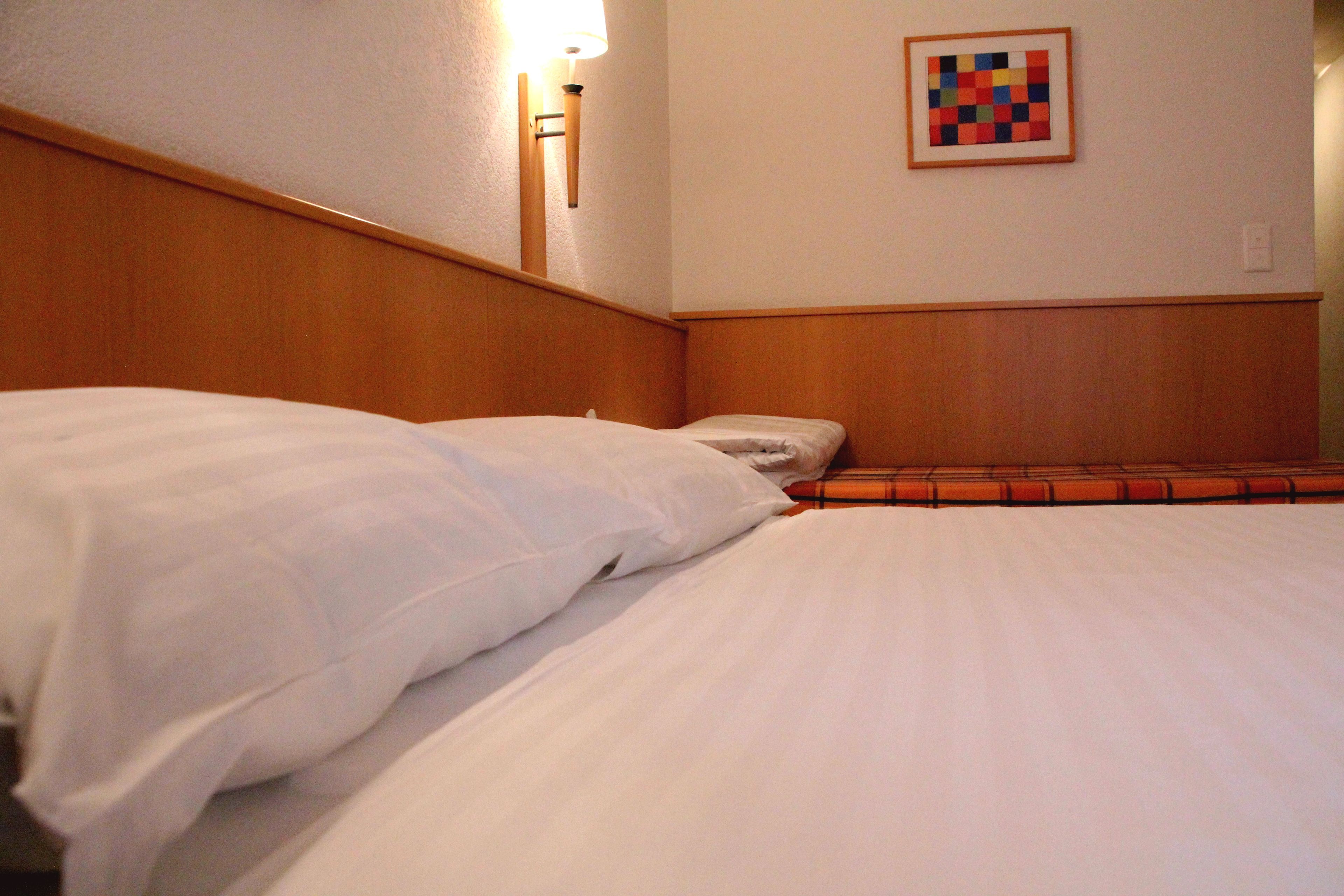 Une chambre d’hôtel avec une tête de lit en bois, un lit recouvert de draps blancs, une lampe fixée au mur et une œuvre d’art encadrée.