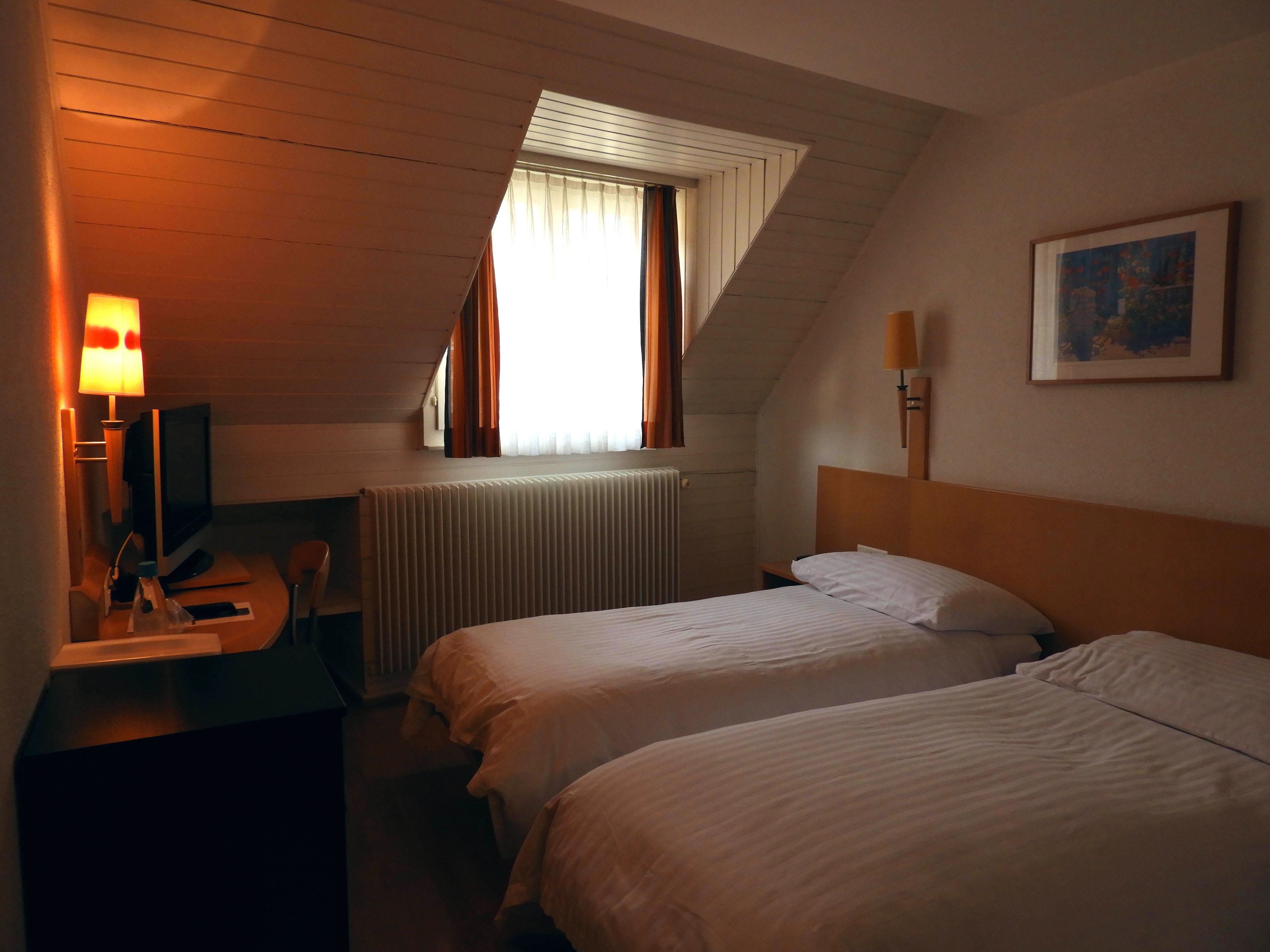 Ein Hotelzimmer mit zwei Betten, einem Schreibtisch mit Computer, einer Lampe, einem Heizkörper und einem Fenster mit Vorhängen.