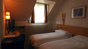 Ein Hotelzimmer mit zwei Betten, einem Schreibtisch mit Computer, einer Lampe, einem Heizkörper und einem Fenster mit Vorhängen.