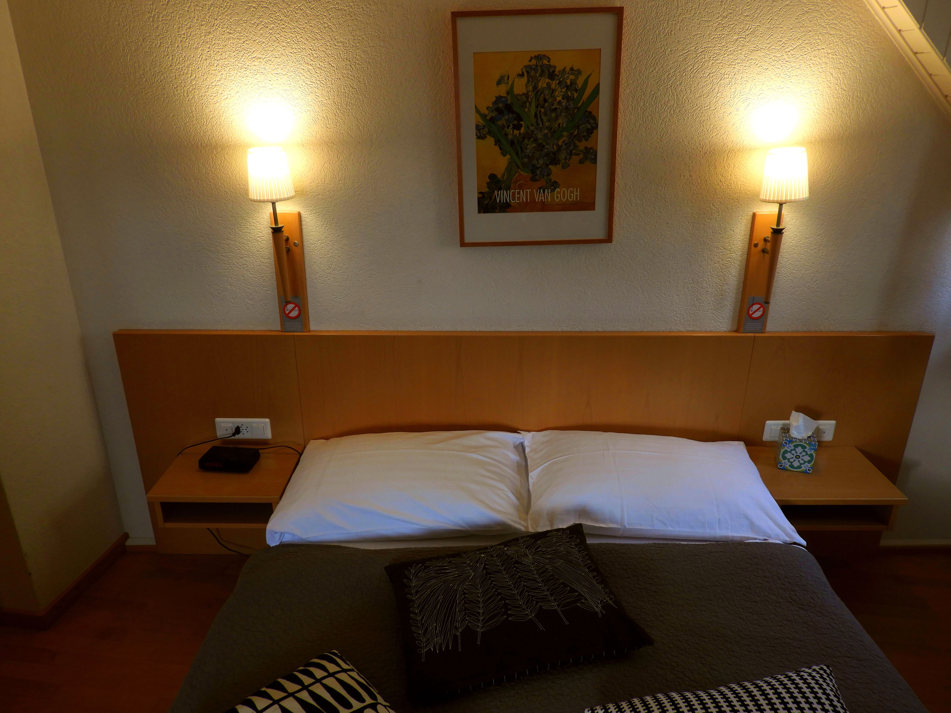 A hotel room with a wooden headboard, a bed with white linens, two bedside tables with lamps, and a framed picture on the wall.