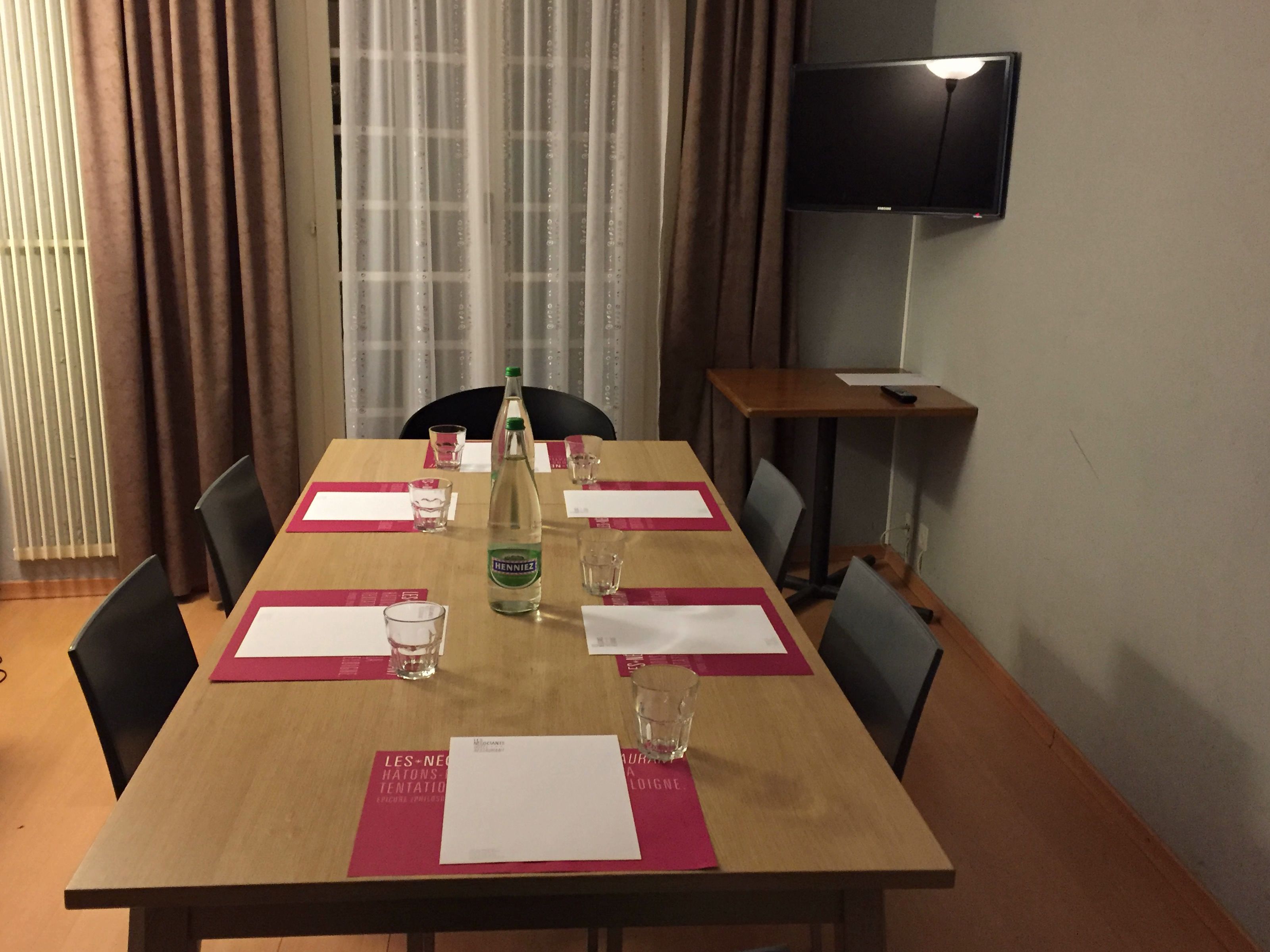 A conference room with a wooden table, chairs, a bottle of Henniez, and a flat-screen TV on the wall.