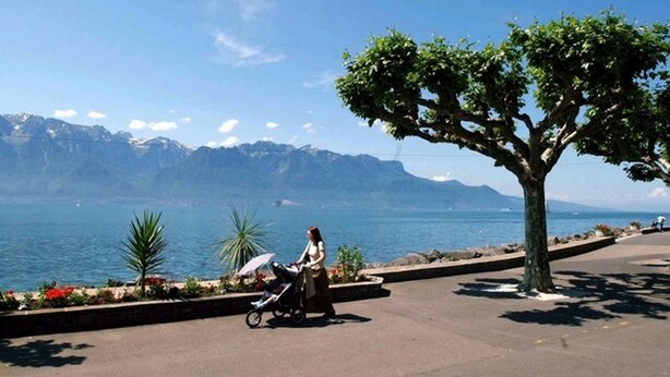 A woman pushing a stroller near a tree-lined waterfront with mountains in the background. - Hotel Restaurant Les Négociants (Vevey)
