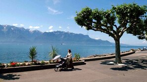 Eine Frau schiebt einen Kinderwagen in der Nähe eines Uferbereichs mit Bäumen, im Hintergrund Berge.