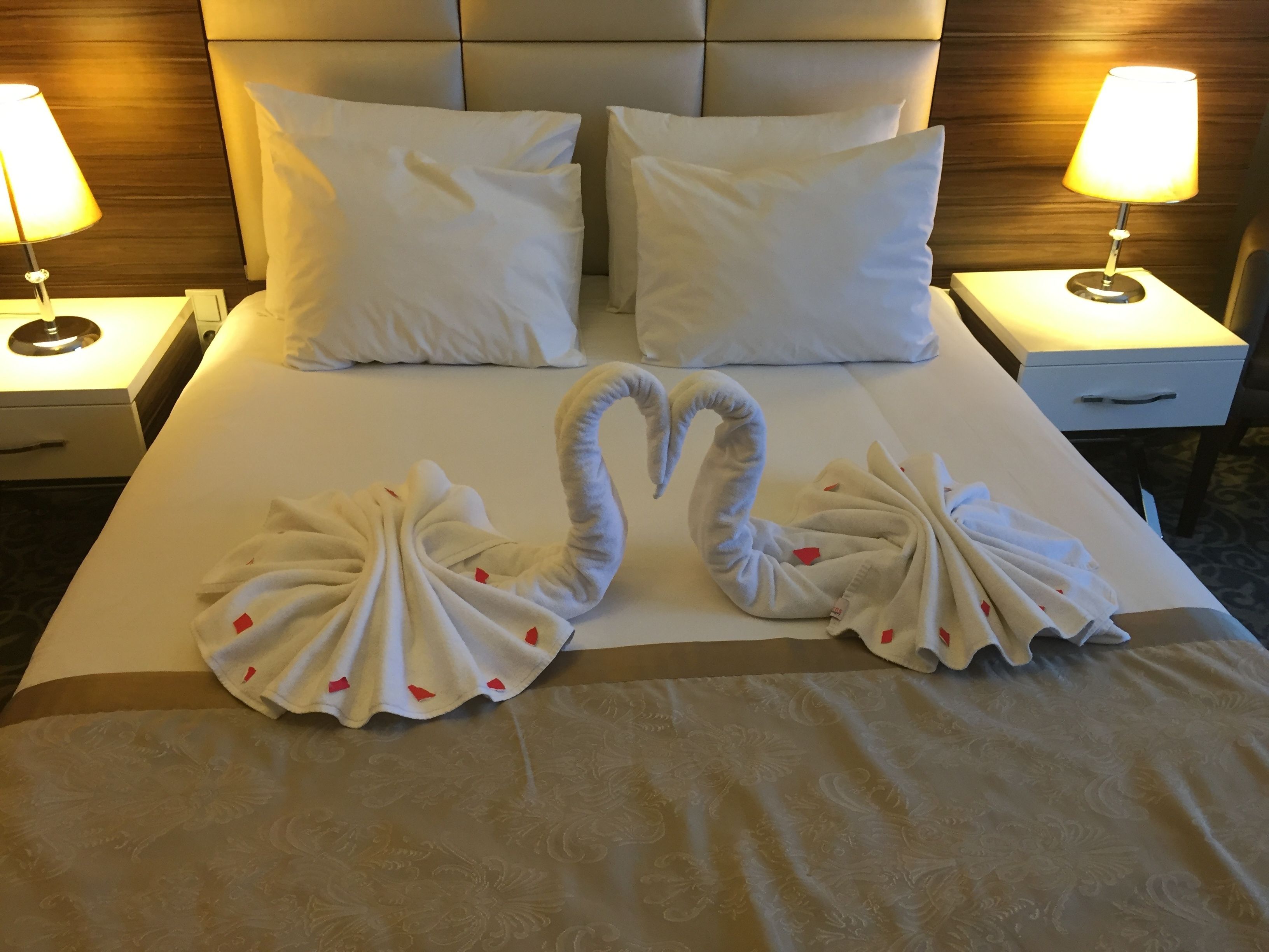 Una camera d'hotel con un letto rifatto, due cuscini, due comodini con lampade e un elegante decorazione a cigno realizzata con asciugamani sul letto.