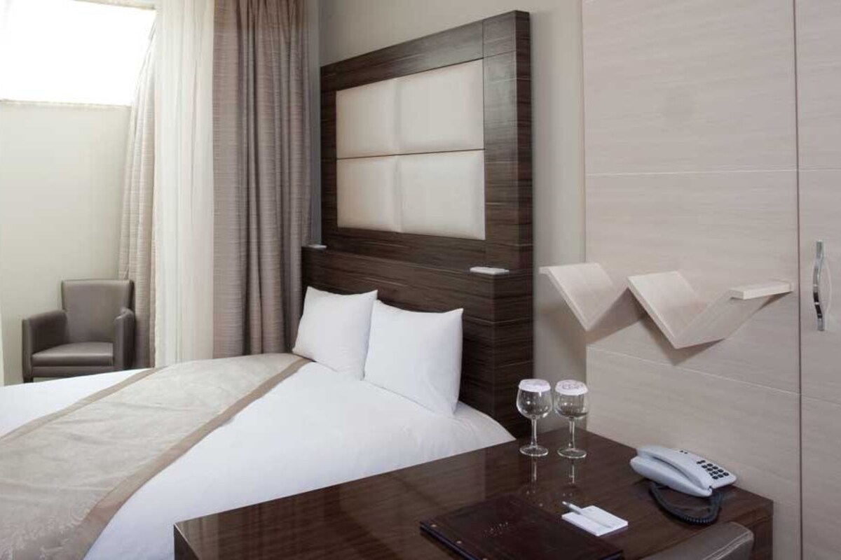 A hotel room with a bed, a bedside table with a phone and glasses, a chair, and a TV mounted on the wall.